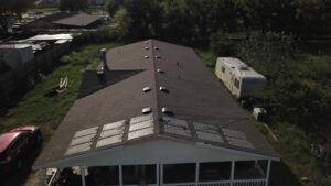 An aerial shot showcasing a long, single-story home with a brand-new dark brown asphalt shingle roof. The roof features numerous evenly-spaced roof vents for proper ventilation and an array of six installed solar panels situated above the covered front porch. The clean, uniform appearance suggests a recent and high-quality professional roof installation.