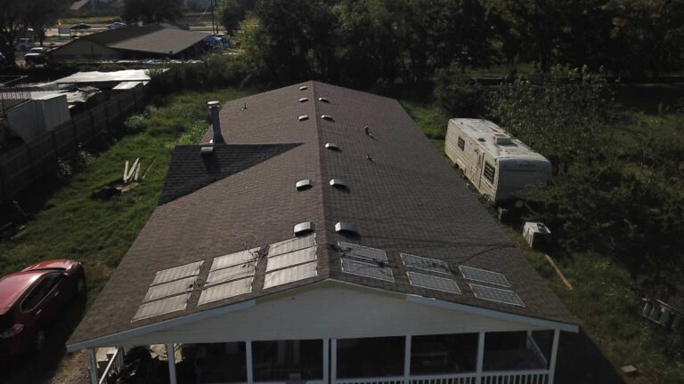 An aerial shot showcasing a long, single-story home with a brand-new dark brown asphalt shingle roof. The roof features numerous evenly-spaced roof vents for proper ventilation and an array of six installed solar panels situated above the covered front porch. The clean, uniform appearance suggests a recent and high-quality professional roof installation.