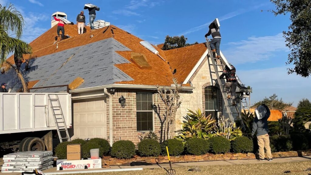 A high-activity residential roofing site showing multiple crew members simultaneously working on a full roof replacement. Workers are seen carrying materials up ladders and installing new brown asphalt shingles and gray underlayment. This image highlights the scale and coordination necessary for a professional project, emphasizing the complexity that amateur work often overlooks, leading to costly roof repair mistakes.