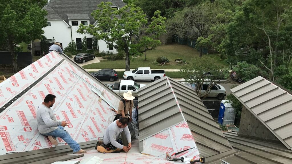 A professional job site view showing the ongoing replacement of a residential roof with standing seam metal panels. Multiple crew members are visible, demonstrating the team effort required for a complex installation. Sections of the roof deck are prepped with Titanium PSU synthetic underlayment, ensuring water resistance and durability beneath the new gray metal panels.