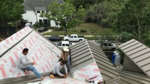 A professional job site view showing the ongoing replacement of a residential roof with standing seam metal panels. Multiple crew members are visible, demonstrating the team effort required for a complex installation. Sections of the roof deck are prepped with Titanium PSU synthetic underlayment, ensuring water resistance and durability beneath the new gray metal panels.