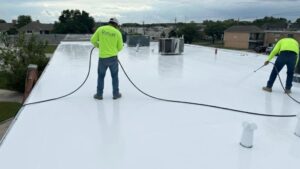 Two roofing contractors in high-visibility shirts using a spray system to apply a white reflective acrylic roof coating on a flat commercial roof.