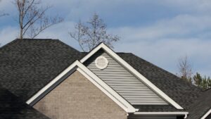 A close-up view of a high-quality asphalt shingle roof replacement featuring dark grey architectural shingles on a multi-gabled house with grey siding and a round white vent.