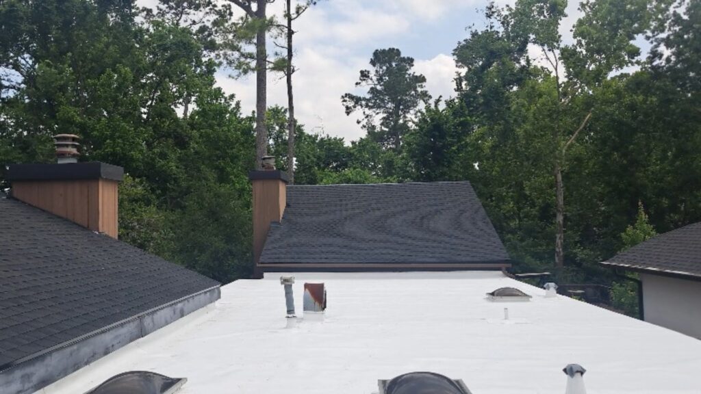 A wide view of a pristine, newly completed bright white commercial TPO flat roof section nestled against dark asphalt shingle roof peaks and surrounded by mature green trees.