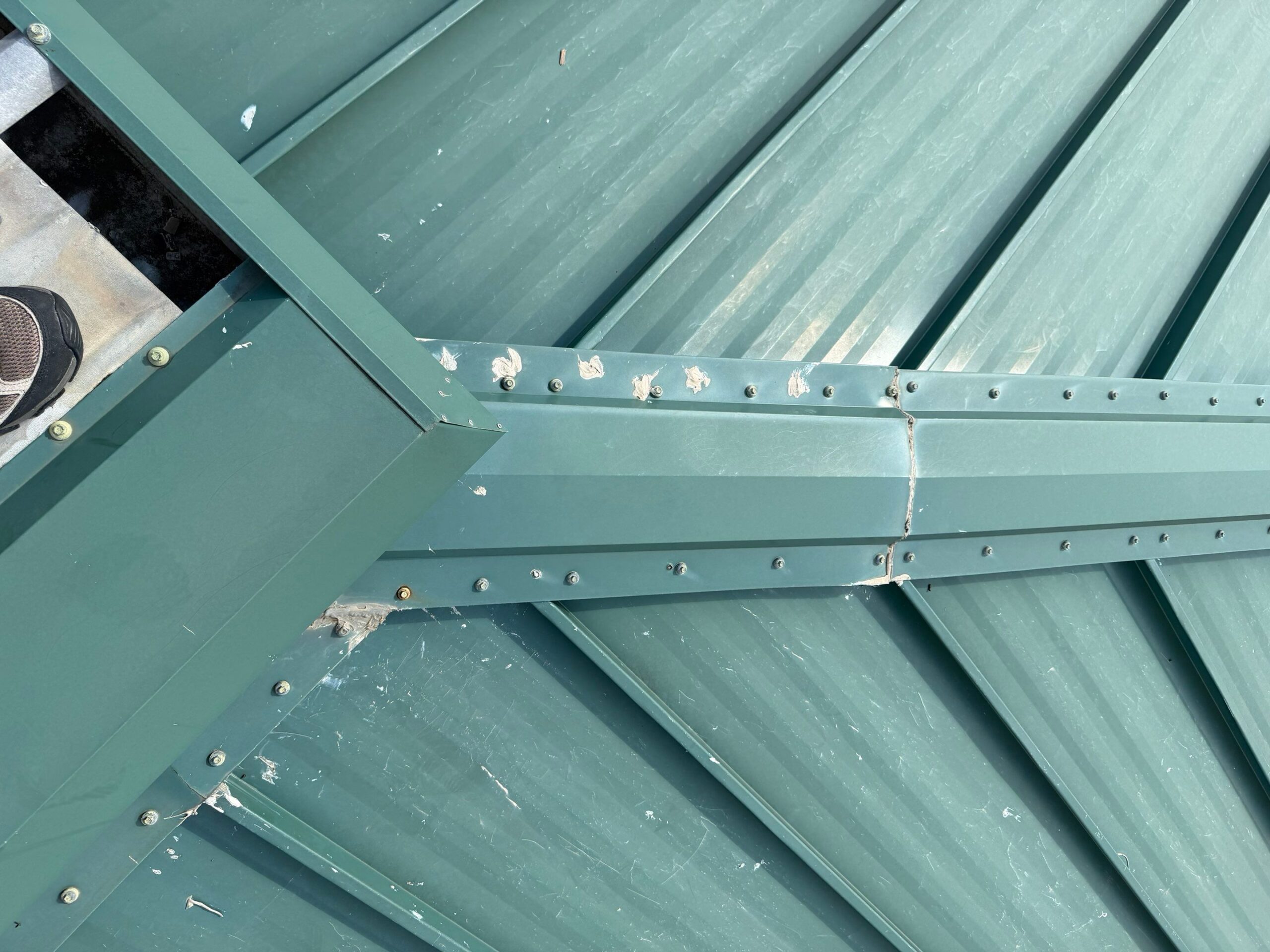Top-down view of valley flashing details, sealant smudges, and fastener joints on a green standing seam metal roof, by Achilles.