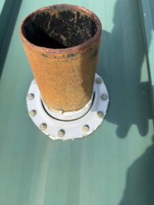 Rusted metal vent pipe penetration sealed with a grey rubber flashing boot on a green standing seam metal roof, by Achilles.